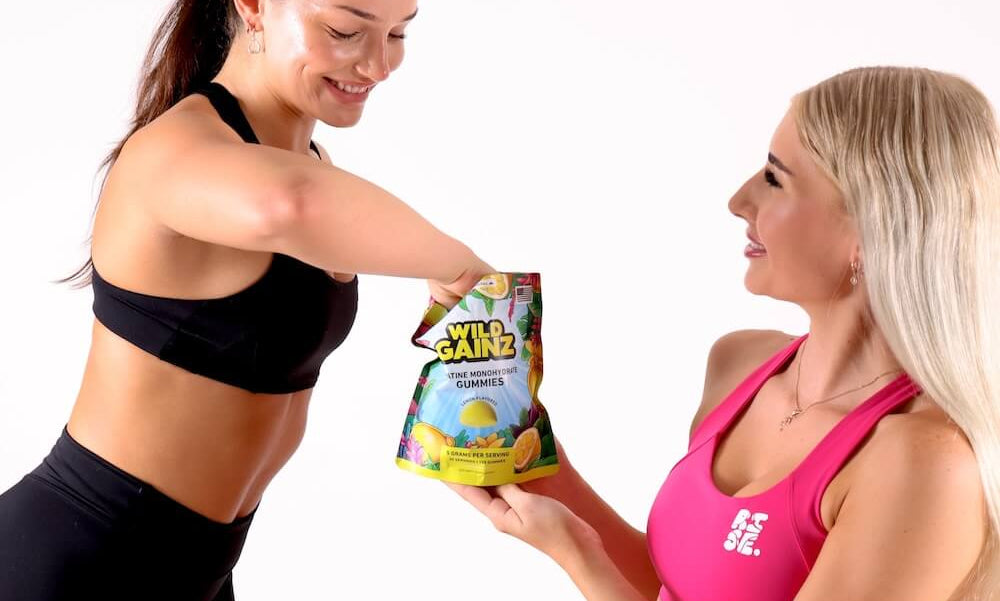 Two women in athletic wear are shown in a studio setting, with one handing a bag of Xtrix Grapefruit Flavored Energy Bites to the other. The woman on the right is wearing a bright pink matching activewear set with the Xtrix logo, and the woman on the left wears a black matching set.